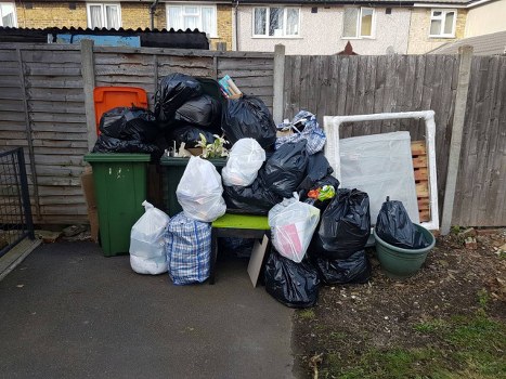 A collection of waste bags and rubbish items piled on a small outdoor paved area next to a wooden fence, with several black plastic rubbish bags, white bin liners, and a few checkered fabric bags containing assorted waste. There is a green wheelie bin and a green plant pot visible among the waste. An orange recycling bin is positioned in the background, and part of a dismantled bed frame or large white flat panel rests against the fence, surrounded by dirt and some sparse patches of grass. The waste appears to be prepared for collection, with the scene set in a residential area possibly in Baldock, Hertfordshire, indicating a typical rubbish disposal site managed by a local waste removal service such as Commercial Waste Baldock.