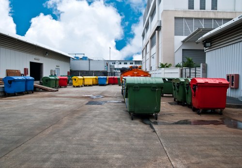 A paved outdoor area adjacent to a commercial building features multiple large wheelie bins in various colours including green, red, blue, yellow, and black, arranged in a row along the open space. The bins are made of durable plastic with textured surfaces and are fitted with metal lids and wheels for mobility. The surrounding environment includes two industrial-style metal structures with ventilation panels on either side, and additional equipment visible in the background. The area is well-lit under a partly cloudy sky, with some shadows cast by the bins and nearby structures. This scene illustrates a typical waste management setup for a commercial premises, commonly handled by local rubbish removal services such as Commercial Waste Baldock, serving the Baldock area and surrounding postcode regions.