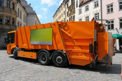 A large, bright orange commercial waste collection vehicle, equipped with multiple storage compartments and a hydraulic lifting mechanism, is parked on a cobblestone street in an urban area. The vehicle features a distinctive green reflective strip on its side for visibility, and its surface appears clean with a smooth, painted metal finish. The street is lined with historic, multi-storey buildings with cream and beige facades, ornate balconies, and tall windows, suggesting a European town centre environment. The background shows a partly cloudy sky with patches of blue, indicating daytime. Surrounding the vehicle, parts of other structures and pavement are visible, but no pedestrians or waste materials are present in the scene. The vehicle is associated with commercial rubbish collection services, possibly operated by Commercial Waste Baldock, serving the local area near the postcode inferred from the Baldock location. The scene captures the typical urban context for rubbish removal operations, emphasizing clean, professional waste management in a city environment.