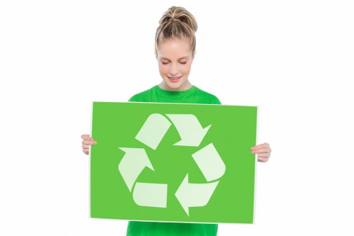 A young woman with blonde hair styled in a high ponytail is smiling and wearing a bright green long-sleeved shirt. She is holding a large, rectangular green sign featuring a white recycling symbol composed of three arrows forming a triangle, indicating recycling and waste management. The background is plain and white, highlighting her and the sign. The image emphasizes environmental awareness and responsible waste disposal, aligning with rubbish removal services such as those offered by Commercial Waste Baldock, serving local communities in the area including the postcode region around Baldock. The woman’s cheerful expression and the prominent recycling symbol support themes of sustainability, waste reduction, and environmentally friendly waste management practices, making the image relevant for businesses involved in rubbish collection and disposal services in the UK.
